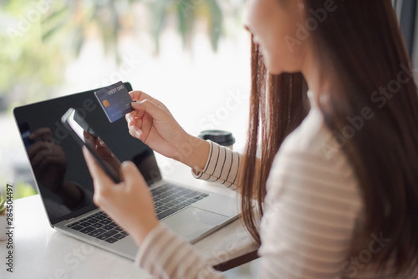 Fototapeta Closed up and focus on hand of young woman hold a credit card and other hand using a smart phone which is shopping online and sitting on table near big mirror at cafe. online payment concept.