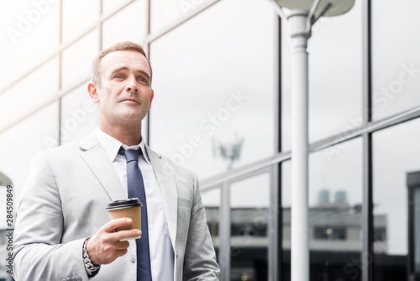 Fototapeta Be ambitious. Businessman in suit is walking along the building with take-out coffee. Copy space on the right side