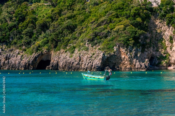 Fototapeta Landscape with turquoise calm sea water, mountain with rocky hillside covered with green trees and bushes and caves, cruise touristic boat. Corfu Island, Greece. 