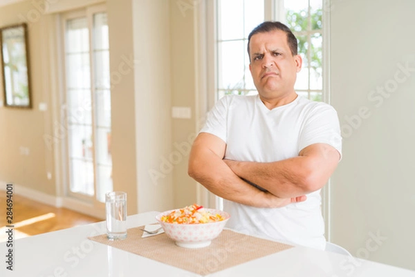 Fototapeta Middle age man eating rice at home skeptic and nervous, disapproving expression on face with crossed arms. Negative person.
