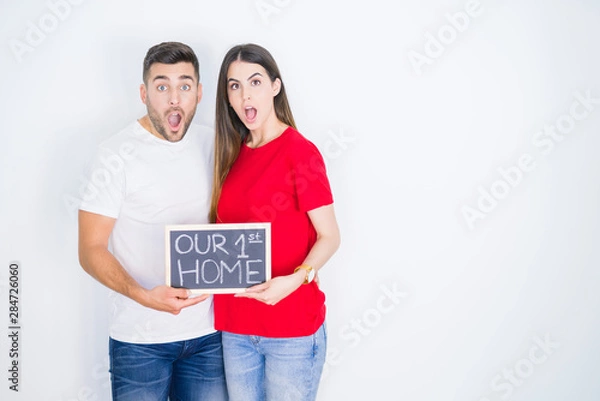 Fototapeta Young beautiful couple holding blackboard with new home text over white isolated background scared in shock with a surprise face, afraid and excited with fear expression
