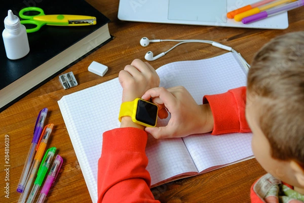 Fototapeta Little boy sitting at the table and looking smart watch. Smart watch for baby safety. The child makes school lessons, listening to music, calling friends.