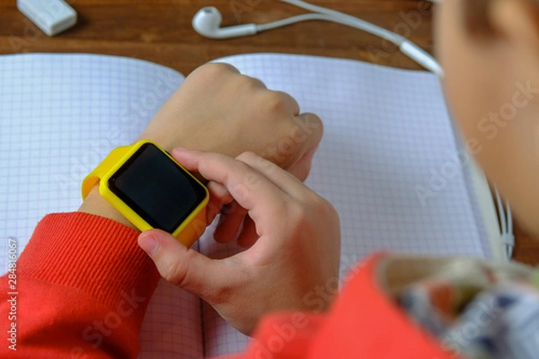 Fototapeta Little boy sitting at the table and looking smart watch. Smart watch for baby safety. The child makes school lessons, listening to music, calling friends.