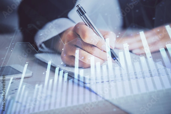 Fototapeta A woman hands writing information about stock market in notepad. Forex chart holograms in front. Concept of research. Multi exposure