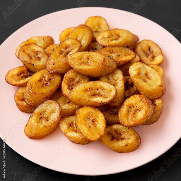 Fototapeta Homemade fried plantains on a pink plate on a black background, side view. Close-up.