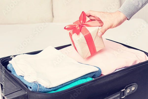 Fototapeta Suitcase on the bed, man will collect clothes for the road and puts the gift with a red ribbon in the suitcase, man's hand, closeup, cropped image, toned