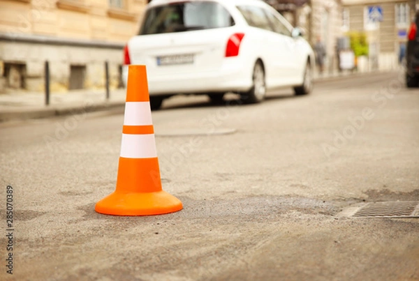 Fototapeta Repair work sign restricting traffic. White-orange plastic striped cone warning of danger. Tightening in traffic. Street of the old city with cars. Roadway repair. Poor asphalt on the road. Attention