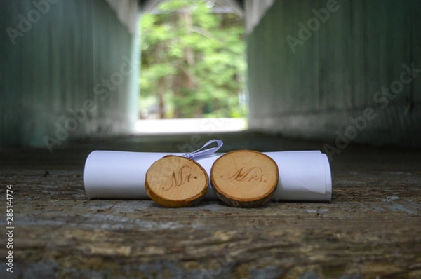 Obraz Rustic Mr. & Mrs. signs cut from tree branches sit in front of a rolled marriage license at the end of a covered wooden bridge