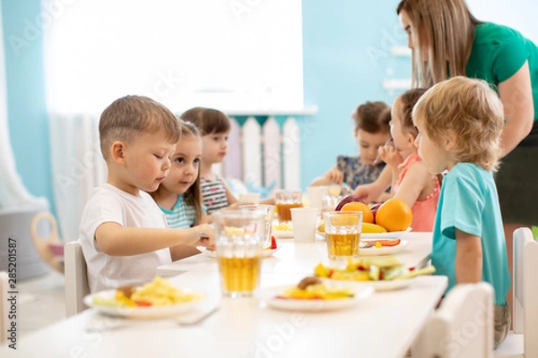 Fototapeta Dzieci jedzą obiad w przedszkolu. Mali chłopcy i dziewczynki z grupy dzieci siedzą przy stole przy obiedzie i jedzą apetycznie. Dzieci z opiekunem w przedszkolu. Dzieci mają zdrowe