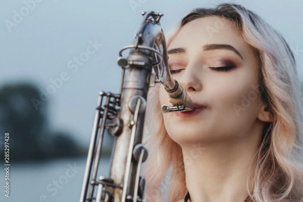 Fototapeta Portrait of young musician on nature background, woman face playing saxophone and blowing into the trumpet, concept music and relax