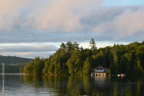 Obraz Boat house on Saranac Lake bathed in the morning sunrise. The coastline is reflected in the water and framed by the colored clouds