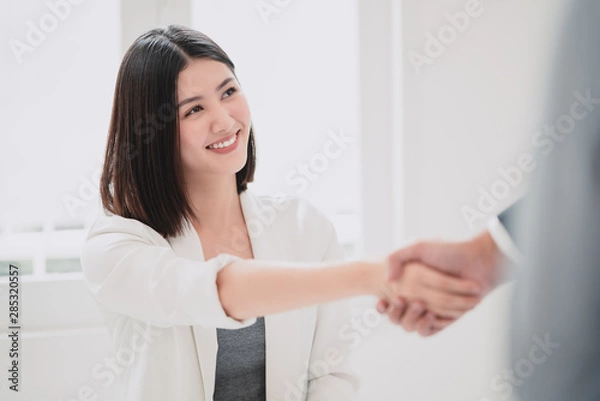 Fototapeta Beautiful young asian business woman smiling with triumphant of the success deal or project while shaking hand with business man with the white natural sunlight as a background.