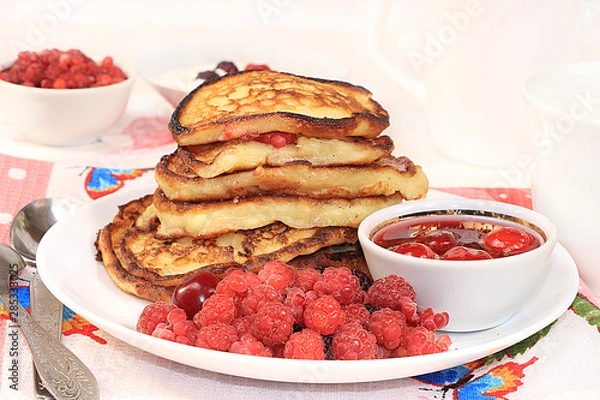 Fototapeta Delicious russian pancakes with cherries and raspberries and a mug of milk on a bright table, selective focus. Traditional Russian pancakes, natural food, jam and berries with milk.