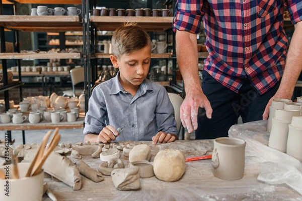 Fototapeta Middle aged caucasian man teaches his little brother to work with clay on pottery workshop. Art work concept.