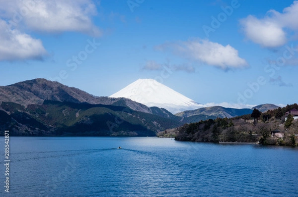 Obraz Mount Fuji in Japan covered by snow seen across a large lake. Small boat leaving a triangle form wake as if mirroring the shape of the mountain. Blue sky, white clouds, spring in coastal forests