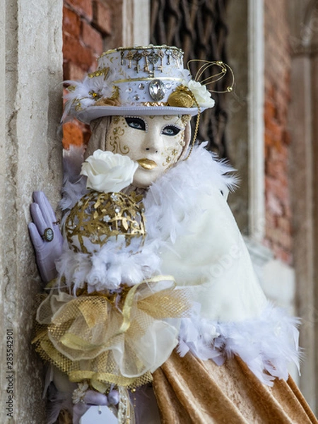 Fototapeta Lady in gold and white costume and masked face at Venice carnival. Highly decorative outfit with shiny details, eyes visible behind the mask. Old palace wall in background