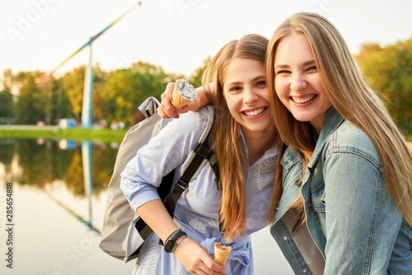 Fototapeta Two pretty student girls, dressed casually, enjoy walking in city amusement park after their college exam, eat ice-cream, pose cheerfully at camera with broad gorgeous smiles, being in joyful mood.