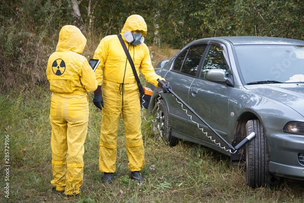 Fototapeta Two dosimetrist  in protective suite and mask with geiger counter measuring radiation level near car