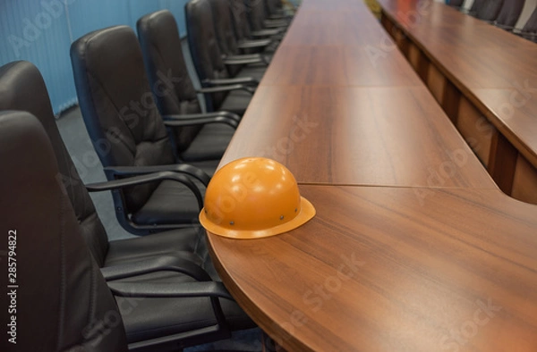Obraz An orange safety helmet left on a long table in a discussion room, with a row of black leather chairs stretching from foreground to background.