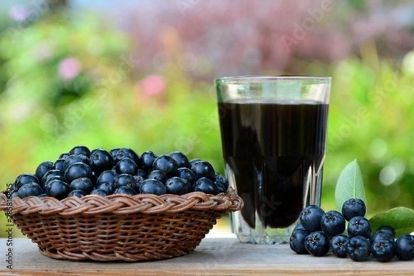 Fototapeta Black chokeberry berries in a basket and a glass of drink on a wooden table on the background of the garden close-up