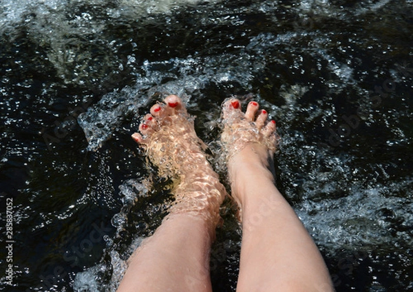 Obraz A pair of feet with red-painted toenails splash in the rushing water of the river.