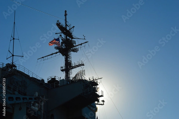Obraz The sun hides behind a United States warship flying the American flag, throwing the ship in shadow and illuminating the flag from behind