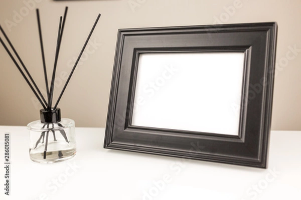 Fototapeta Aromatizer or perfume stick. Aromatic air freshener in a transparent glass bottle with black reeds on a white table and black desk photo frame with a blank space on a white table