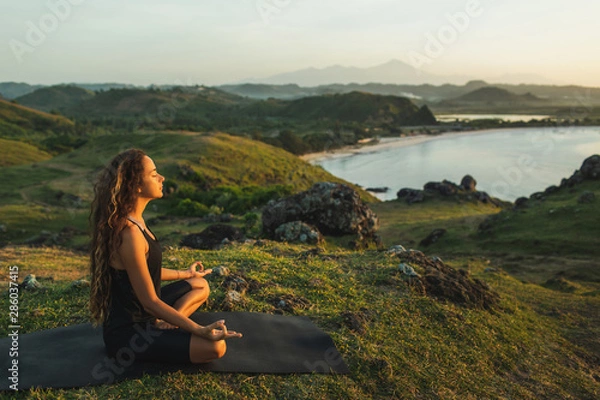 Fototapeta Woman doing yoga alone at sunrise with mountain and ocean view. Harmony with nature. Self-analysis and soul-searching