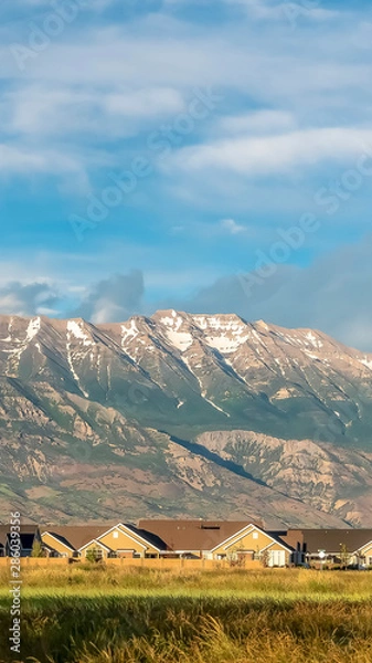 Fototapeta Vertical frame Magnificent mountain towering over homes and vast grassy terrain on a sunny day