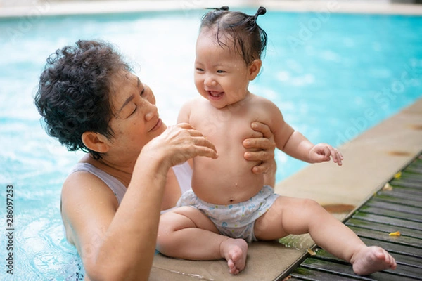 Fototapeta Senior asian woman is enjoying time with her adorable one year old grandchild in a swimming pool.
