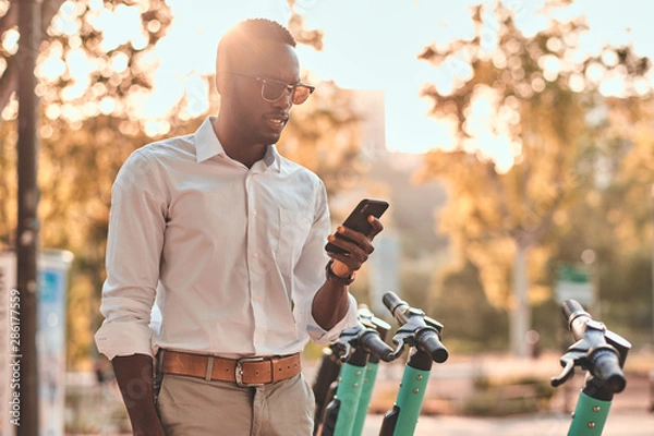 Fototapeta Young african guy is paying for electrical scooter by web payment using his mobile phone.
