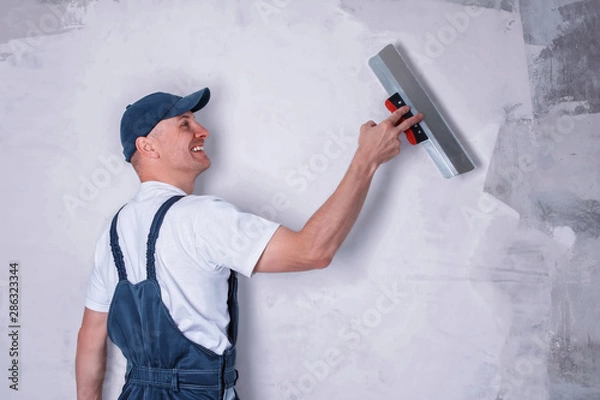 Fototapeta Smiling worker in profile wearing overalls and cap plastering a wall with finishing putty using a putty knife. Repair work and construction concept