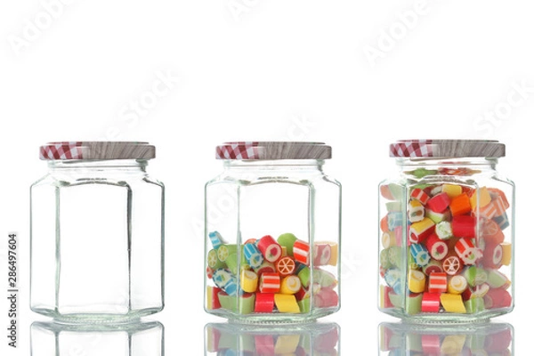 Fototapeta three glass jar, one empty, one half empty and one full of colored candies isolated on white background with clipping path and copy space for your text