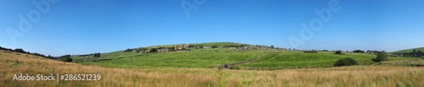 Fototapeta long panoramic view of green fields and village houses and ancient packhorse bridge in rolling west yorkshire dales countryside in colden with blue summer sky