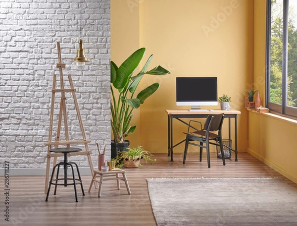 Fototapeta Modern yellow wall background and white brick wall style, wooden working table computer style, easel painting decoration vase of plant.