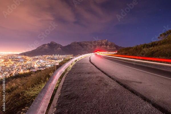 Obraz Long exposure of car driving around a bend with Table Mountain in the background in Cape Town as seen from Signal hill.