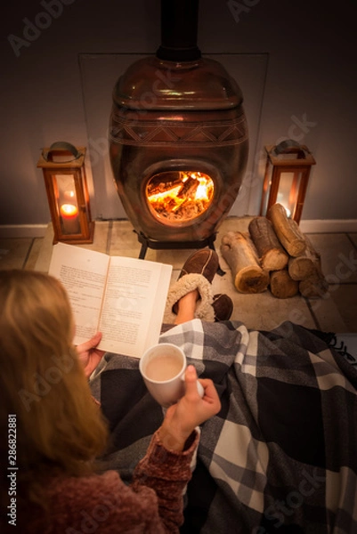 Obraz Woman/girl sitting in front of a cozy fireplace during winter under a blanket  reading a book drinking coffee/hot chocolate.