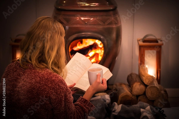 Obraz Woman/girl sitting in front of a cozy fireplace during winter under a blanket  reading a book drinking coffee/hot chocolate.