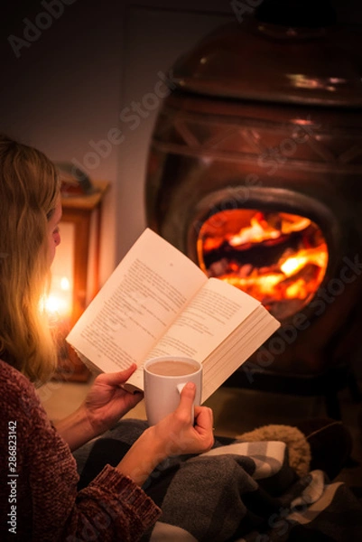 Obraz Woman/girl sitting in front of a cozy fireplace during winter under a blanket  reading a book drinking coffee/hot chocolate.