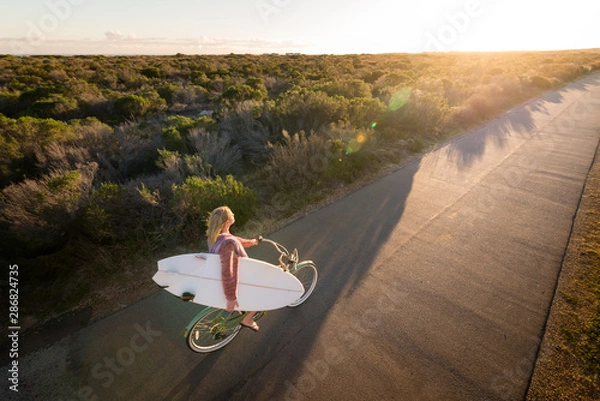 Obraz Beautiful blonde surfer girl on her way to the beach on her bicycle with her surfboard.