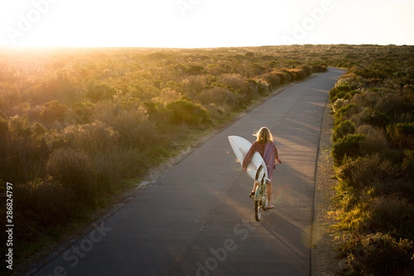 Obraz Beautiful blonde surfer girl on her way to the beach on her bicycle with her surfboard.