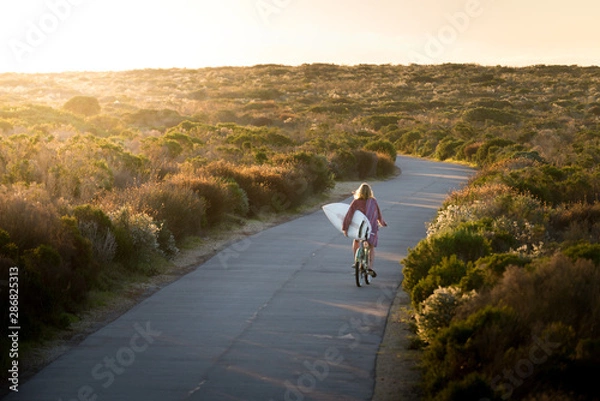 Obraz Beautiful blonde surfer girl on her way to the beach on her bicycle with her surfboard.