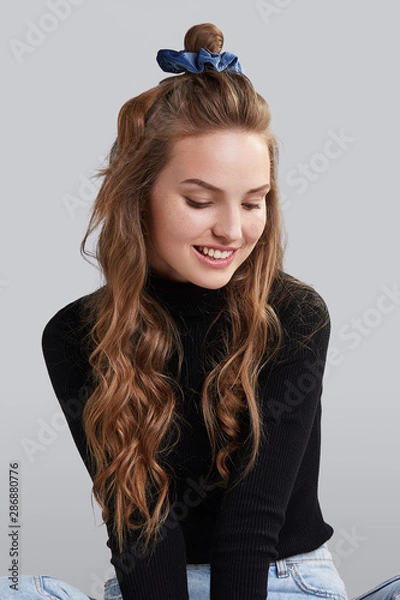 Fototapeta Cropped shot of a ginger girl with wavy hair, sitting on a floor. She wearing black turtleneck and blue jeans. Girl has blue hairpin on her head. Her hair is pulling in high side bun. 
