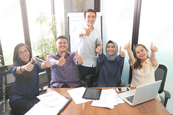 Fototapeta Young creative employees posing on camera in conference room. Company colleagues ready for open brainstorming session, group of multi ethnic business workers smiling at camera, multiple ethnic group