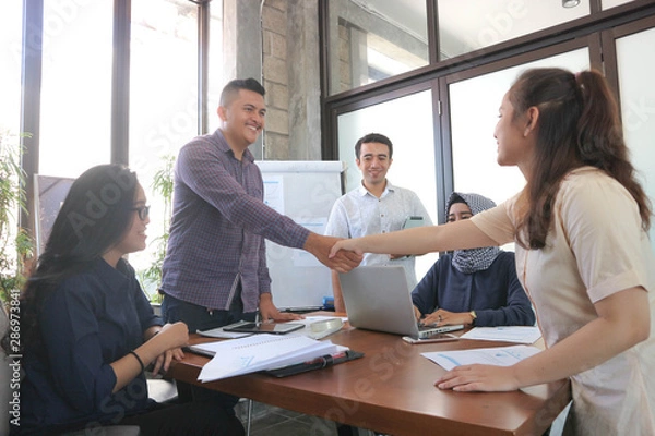 Fototapeta Welcome to team! Two shaking hands and looking at each other with smile while their coworkers sitting at the business meeting