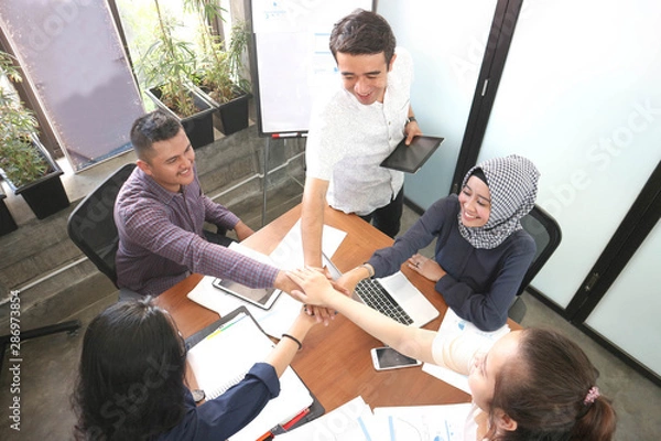 Fototapeta business team giving highfive together at the office meeting room with laptop smartphone and tablet near windows with white board and black board showing cart while looking at camera
