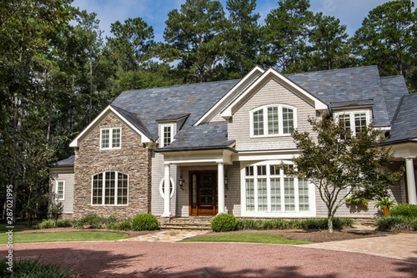 Fototapeta Front view of large estate home in the south with a gravel driveway and lots of windows. house made of brick, stone and clapboard in a cape cod style. and a triple garage with curb appeal