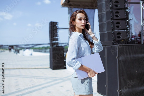 Fototapeta Installation of stage equipment and preparing for a live concert open air. Event manager portrait. Summer music city festival. Young serious woman stand and work with her laptop near the stage...