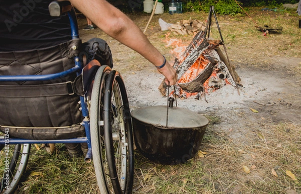 Fototapeta Disabled man resting in a campsite with friends. Wheelchair in the forest on the background of bonfire. Barbeque. Camping.