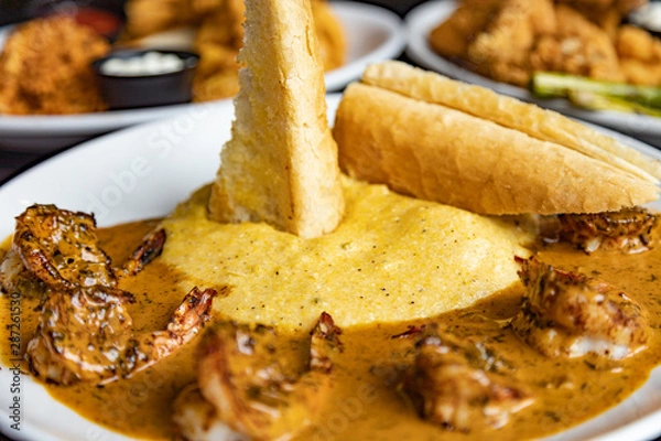 Fototapeta Crispy bread stuck in the grits and shrimp creole cuisine on a white plate. Close up with other platters blurred in the background